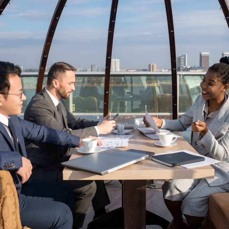 Happy young African businesswoman with paper talking to Chinese colleague at meeting while sitting by table in cafe
